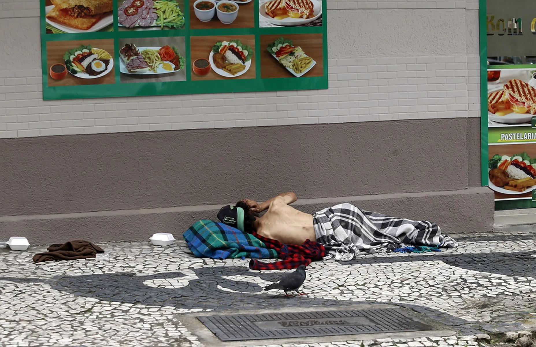 Imagem mostra homem dormindo na calçada, em frente a uma lanchonete, com pombos ao redor