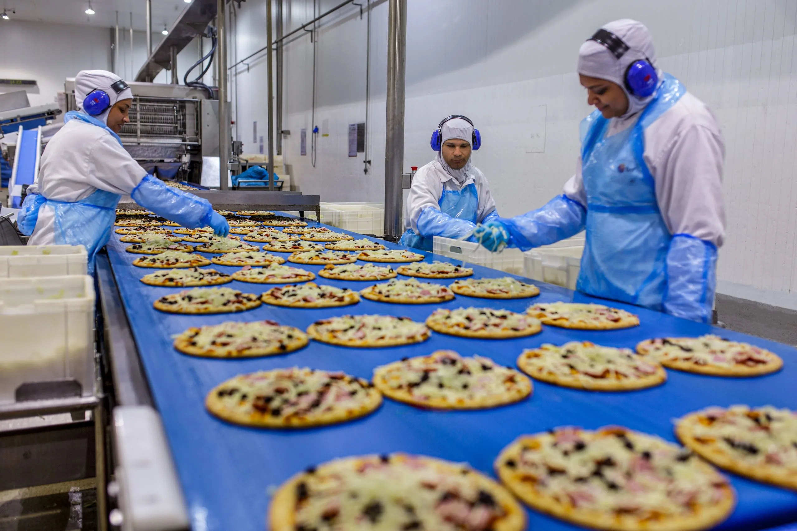 Fábrica de pizzas, mesa cheia de pizzas e três funcionários colocando os ingredientes nas pizzas / maior fábrica de pizzas do Brasil