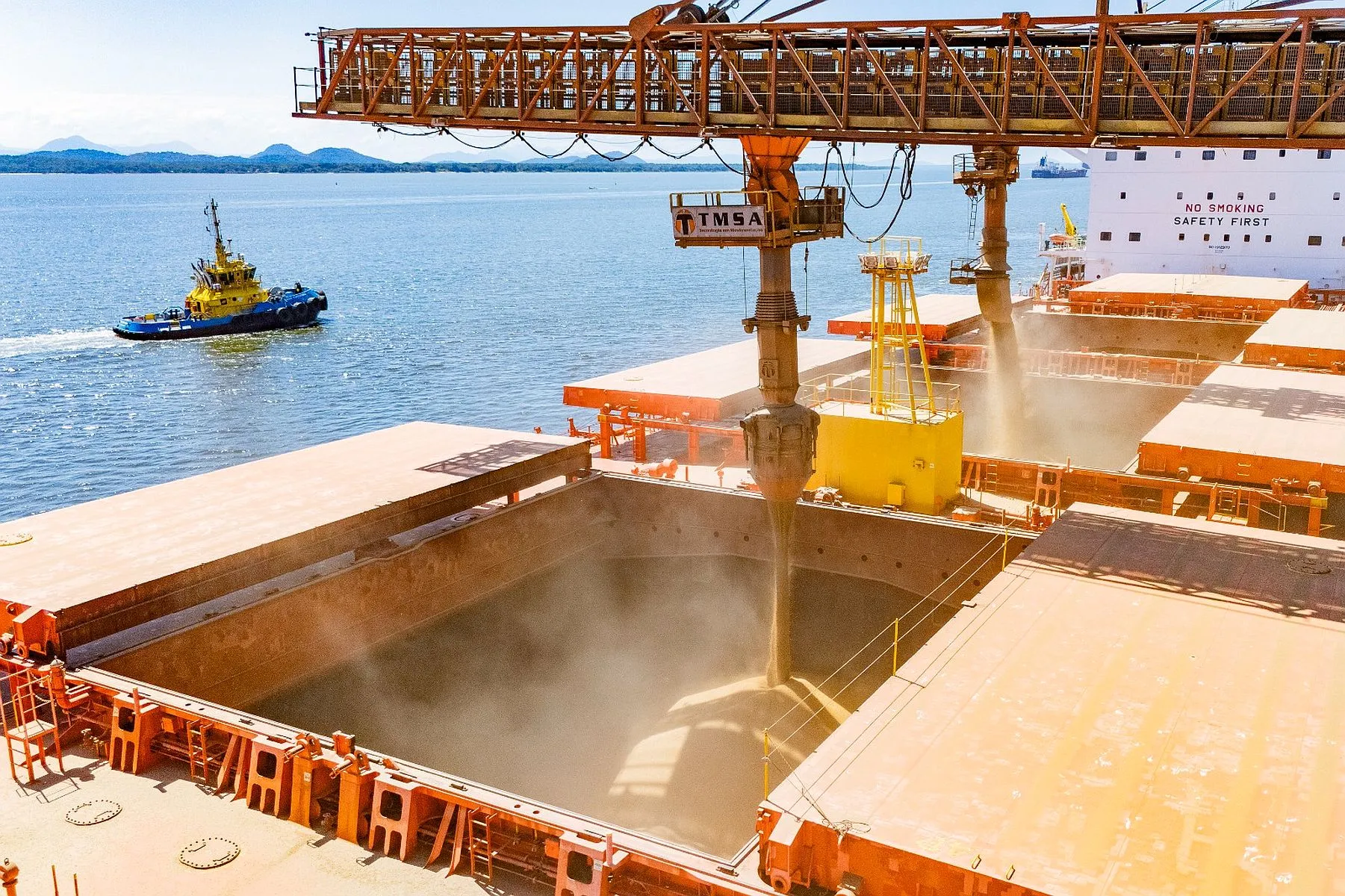 Imagem mostra carregamento de soja em depósito de navio atracado na baía de paranaguá. Ao fundo, um barco pequeno e mais ao fundo as ilhas próximas