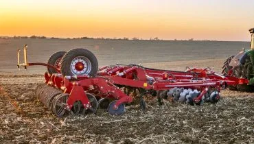 Imagem mostra um maquinário agrícola puxado por um trator em um campo, com por no sol no céu