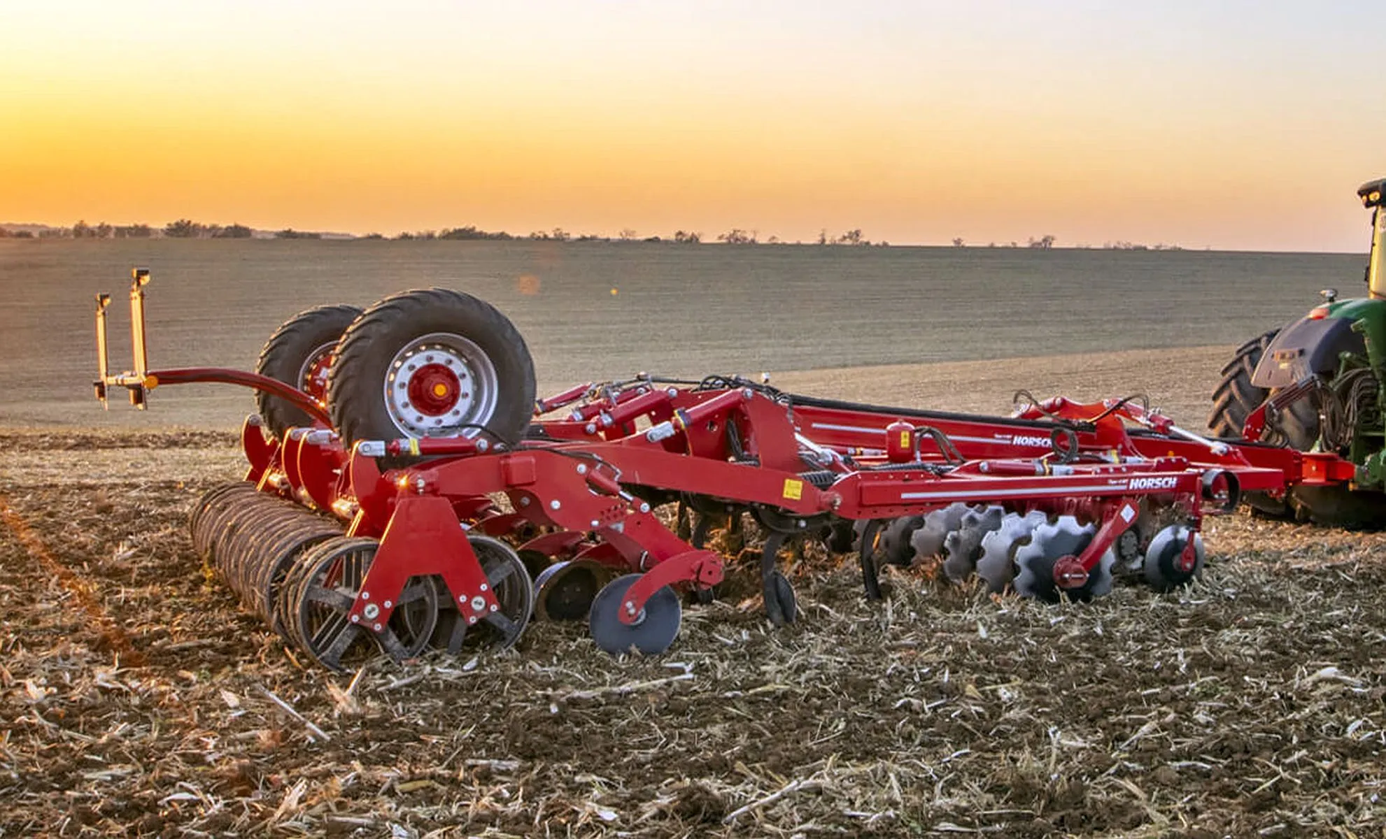 Imagem mostra um maquinário agrícola puxado por um trator em um campo, com por no sol no céu