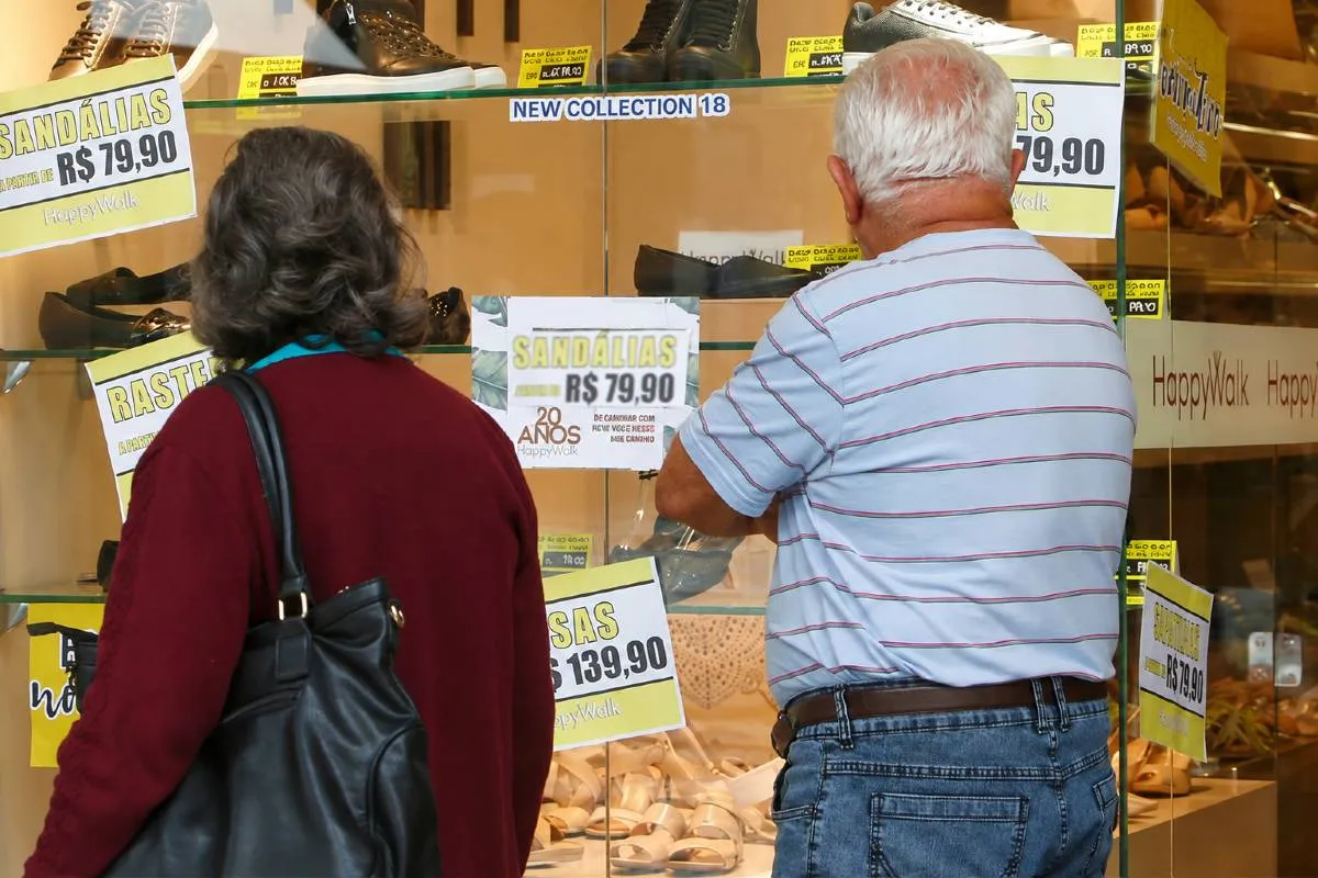 Imagem mostra um casal olhando a vitrine de uma loja com promoções. A placa em detaque diz: Sandálias por R$ 79,90. O casal está de costas, a mulher com uma blusa vermelha, uma bolsa preta e cabelo preso. O homem tem cabelos brancos, usa uma camisa branca com listras pretas e calça jeans. Ele está de braços cruzados olhando os preços desconfiado.