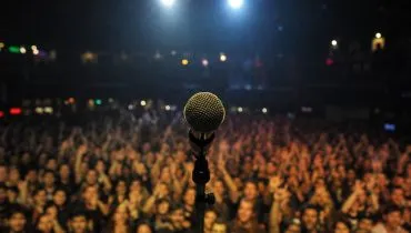Imagem mostra um microfone em um palco com o público ao fundo.