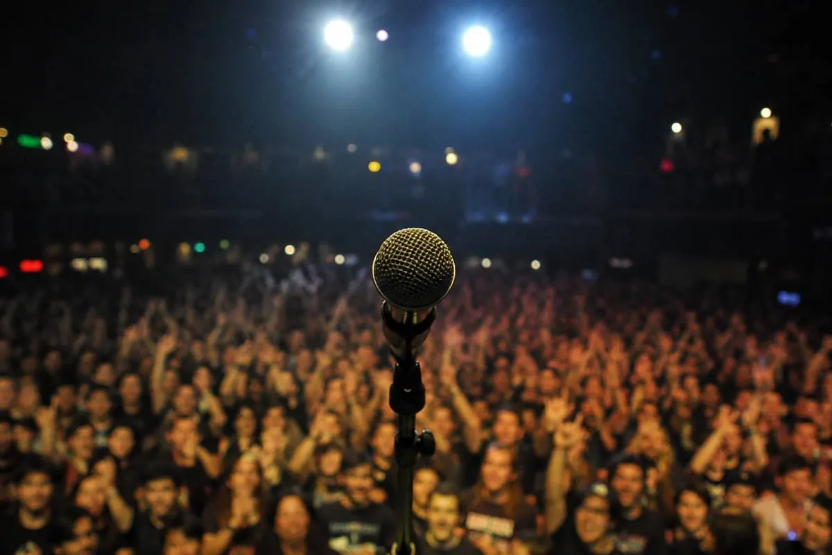 Imagem mostra um microfone em um palco com o público ao fundo.