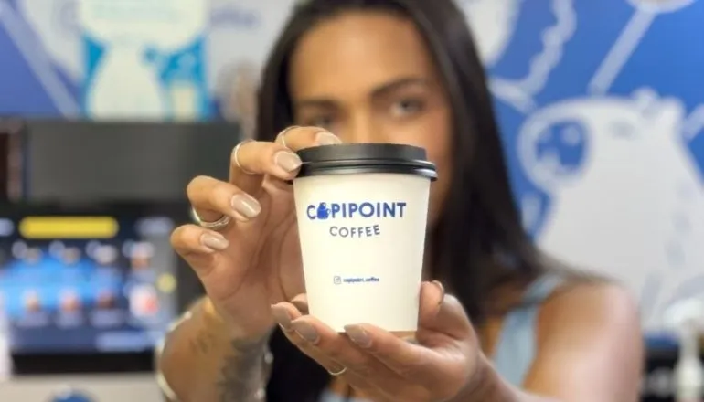 Moça de cabelos pretos lisos segurando um copo de café a sua frente da marca Capipoint / Cafeteria autônoma