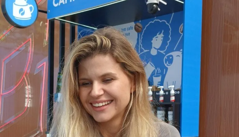 Moça loira de cabelos soltos, sorrindo, em frente ao computador e com una cafeteria autônoma Capipoint atrás dela.