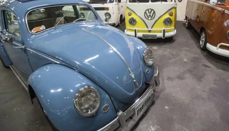 Imagem mostra um fusca azul ao lado de uma kombi amarela e branca.