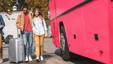 Imagem mostra um detalhe de um ônibus rosa com um jovem casal ao lado com uma mala de viagem.