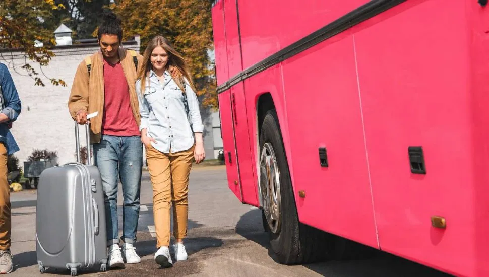 Imagem mostra um detalhe de um ônibus rosa com um jovem casal ao lado com uma mala de viagem.