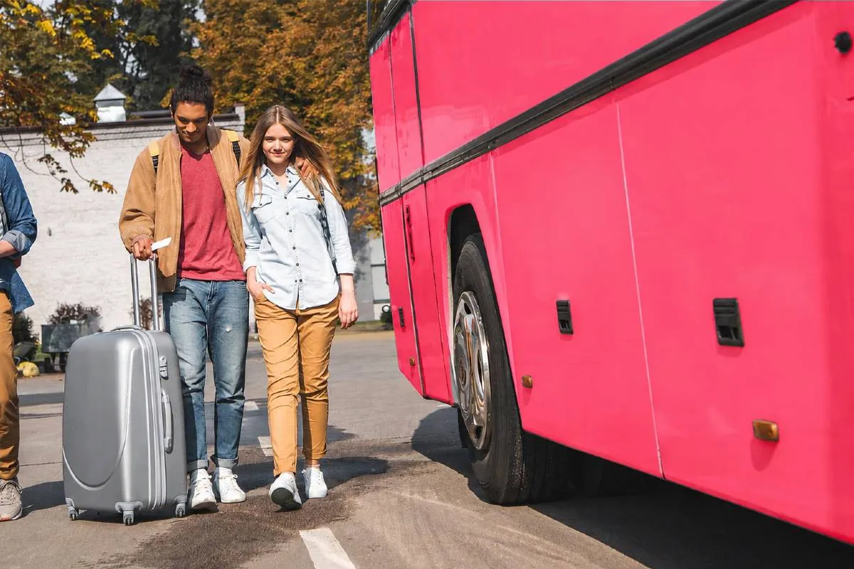 Imagem mostra um detalhe de um ônibus rosa com um jovem casal ao lado com uma mala de viagem.