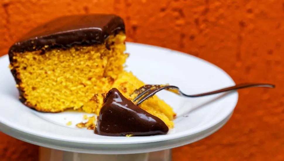 Imagem mostra uma fatia de um bolo de cenoura com chocolate disposta em um prato branco em um fundo laranjado.