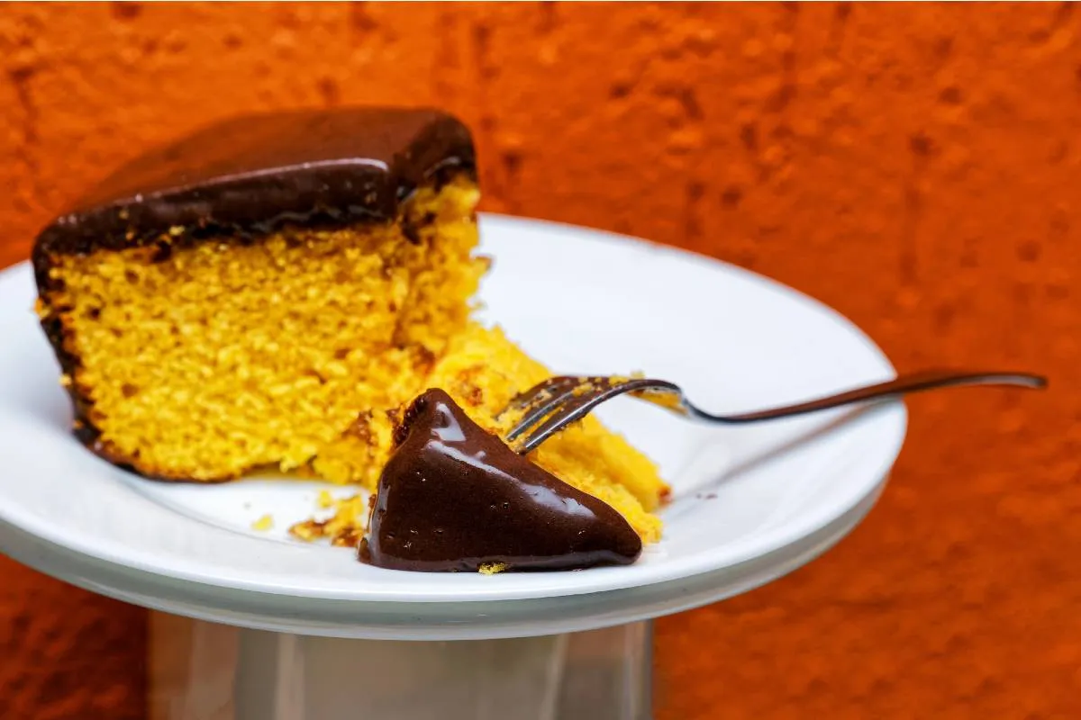Imagem mostra uma fatia de um bolo de cenoura com chocolate disposta em um prato branco em um fundo laranjado.