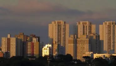 Imagem mostra uma sequência de prédios com nuvens carregadas ao fundo.