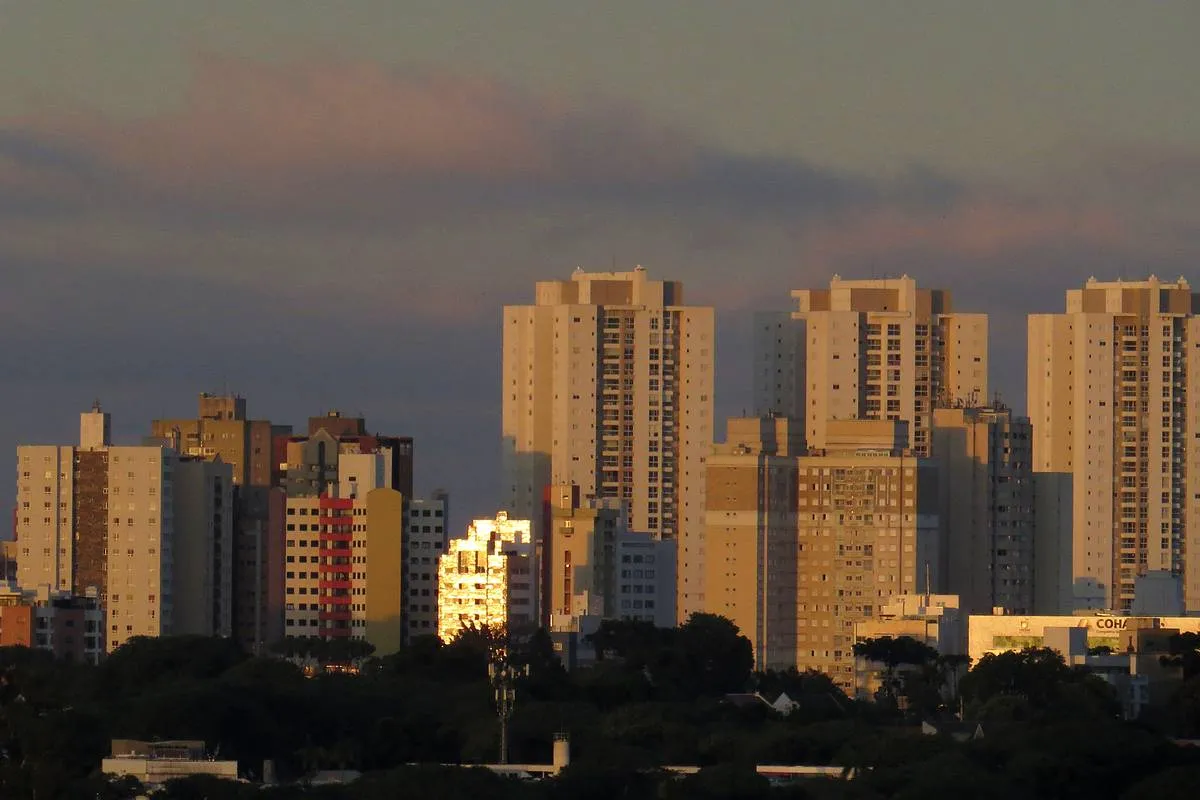 Imagem mostra uma sequência de prédios com nuvens carregadas ao fundo.