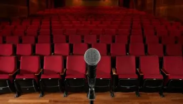 Imagem mostra um teatro vazio ao fundo com um microfone em primeiro plano.