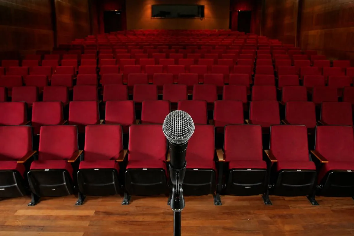 Imagem mostra um teatro vazio ao fundo com um microfone em primeiro plano.