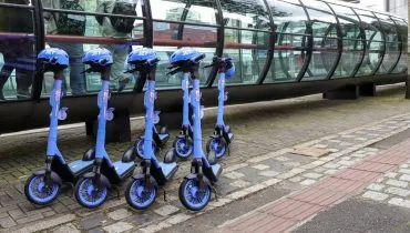 Imagem mostra patinetes azuis na frente de uma estação tubo em Curitiba.