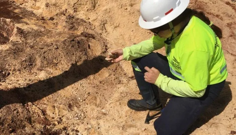 Paleontóloga de capacete e colete analisando amostra de solo em área de escavação.