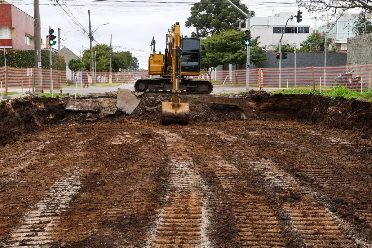 Imagem mostra uma rua em obras, com um grande buraco e uma máquina trabalhando.