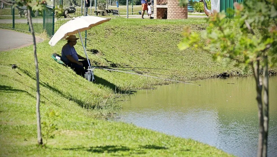 Foto mostra homem sentado à beira de um lado, debaixo de um guarda-sol, pescando