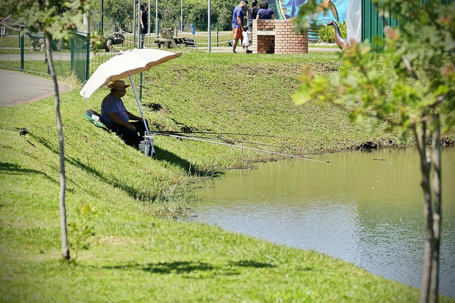 Foto mostra homem sentado à beira de um lado, debaixo de um guarda-sol, pescando