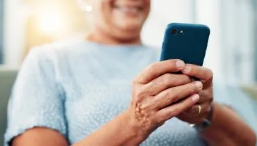Imagem mostra uma mulher com um celular na mão. Ela está de blusa azul clara e o celular tem uma capinha azul escuro.