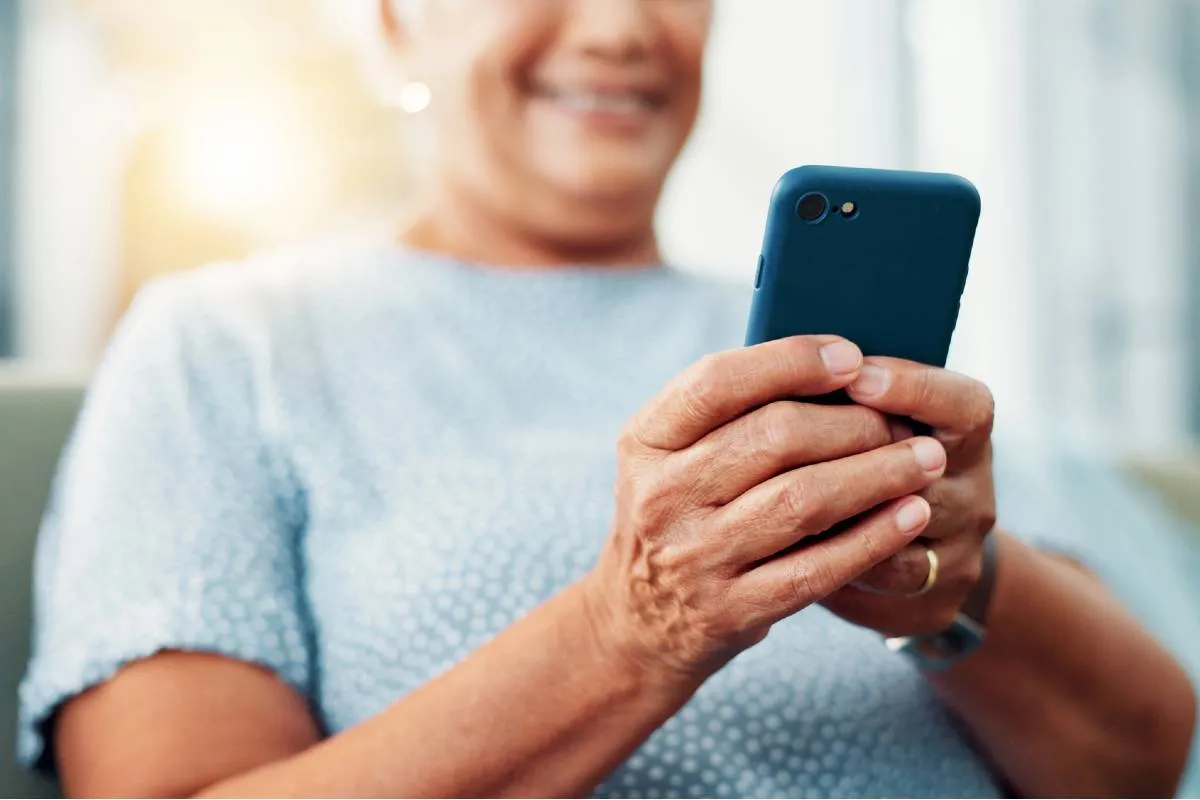 Imagem mostra uma mulher com um celular na mão. Ela está de blusa azul clara e o celular tem uma capinha azul escuro.