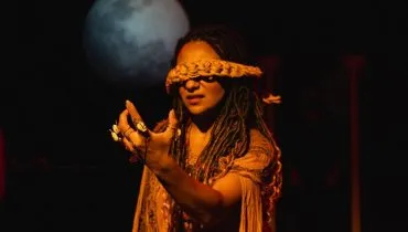 Imagem mostra parte da peça "Ainda Antígona", em cartaz em Curitiba. Na imagem uma mulher vendada com a mão erguida e a lua ao fundo.
