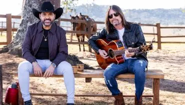 Imagem mostra a dupla sertaneja Edson e Hudson em um sítio. Eles estão sentados em um banco com um cavalo ao fundo. Um dos integrandes da dupla tem cabelo comprido, óculos e segura um violão. O outro está com um chapeu preto.