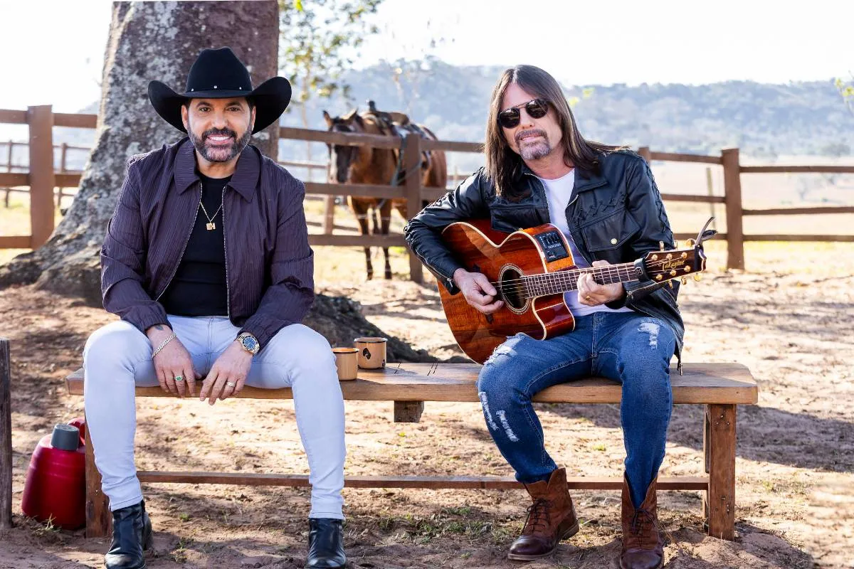 Imagem mostra a dupla sertaneja Edson e Hudson em um sítio. Eles estão sentados em um banco com um cavalo ao fundo. Um dos integrandes da dupla tem cabelo comprido, óculos e segura um violão. O outro está com um chapeu preto.