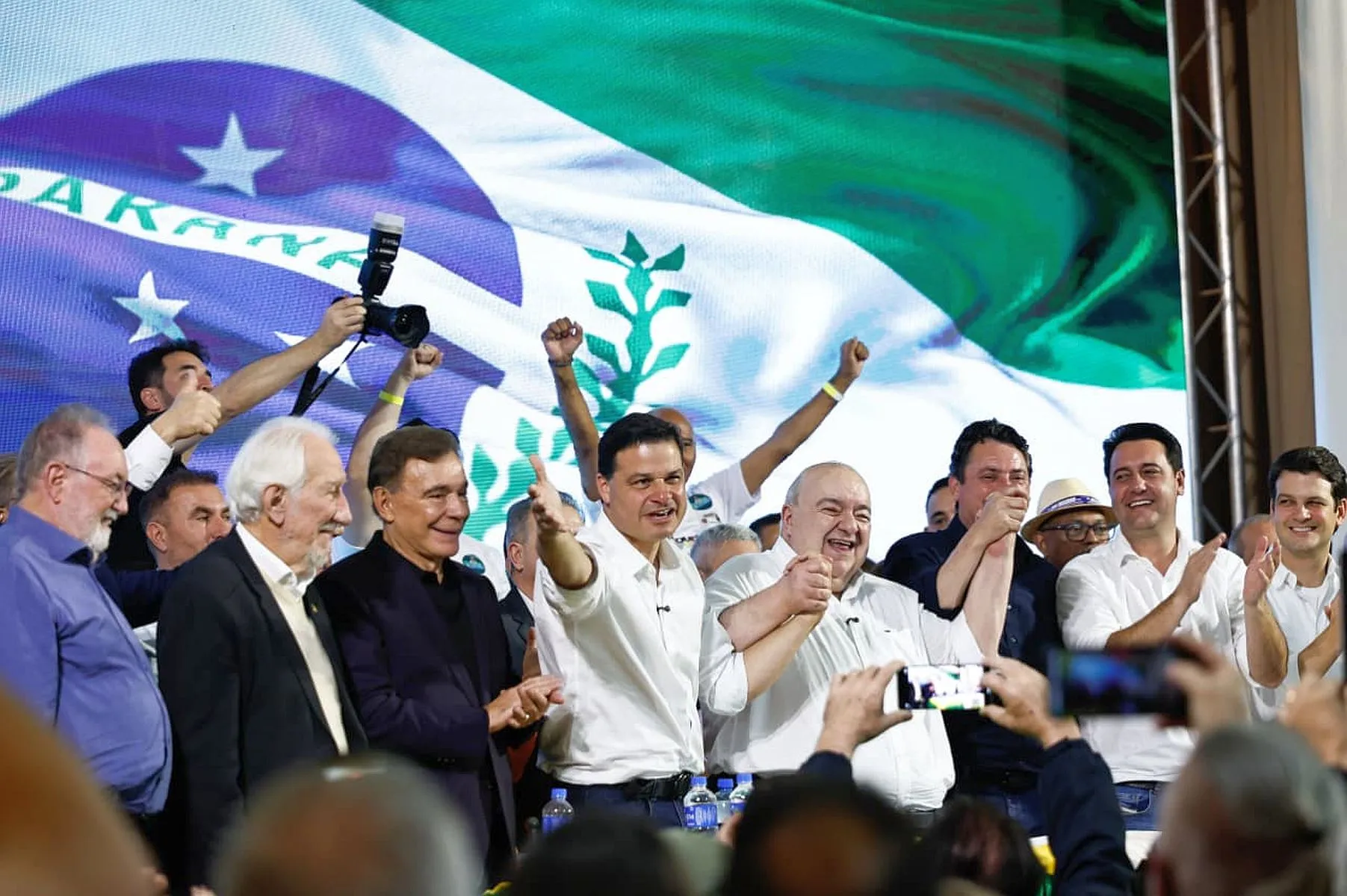 foto mostra diversas lideranças, entre eles o governador Ratinho Junior e o prefeito de Curitiba, Eduardo Pimentel, junto com Sandro Alex, Rafael Greca e Álvaro Dias em cima de um palco, com a bandeira do Paraná num telão ao fundo