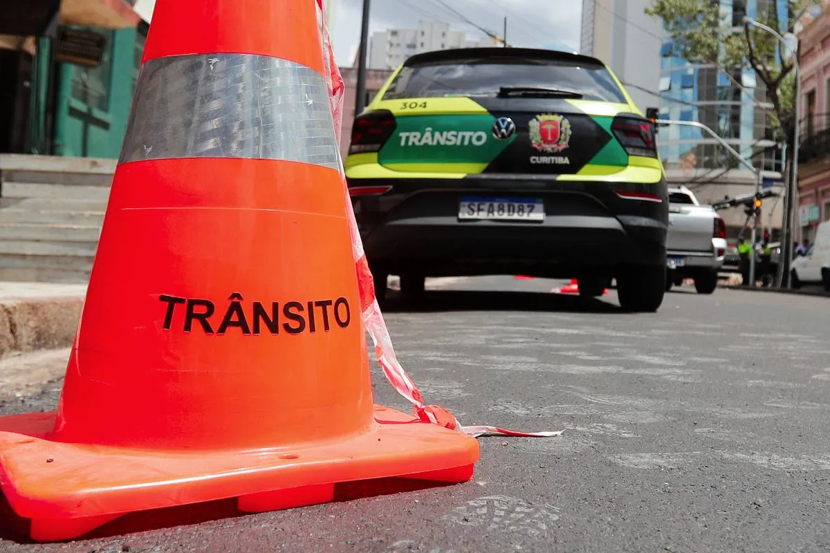 Imagem mostra um cone com o escrito "Trânsito" e uma viatura da Setran ao fundo. A viatura tem as cores verde limão e preto.