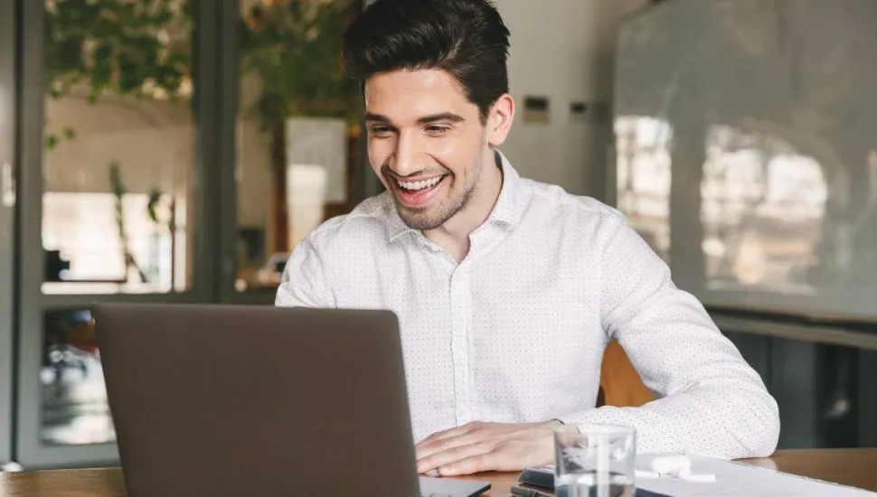 Imagem mostra um homem usando uma camisa branca em frente a um notebook. Ele está olhando para o computador e rindo. Tem um copo de água no canto da mesa.