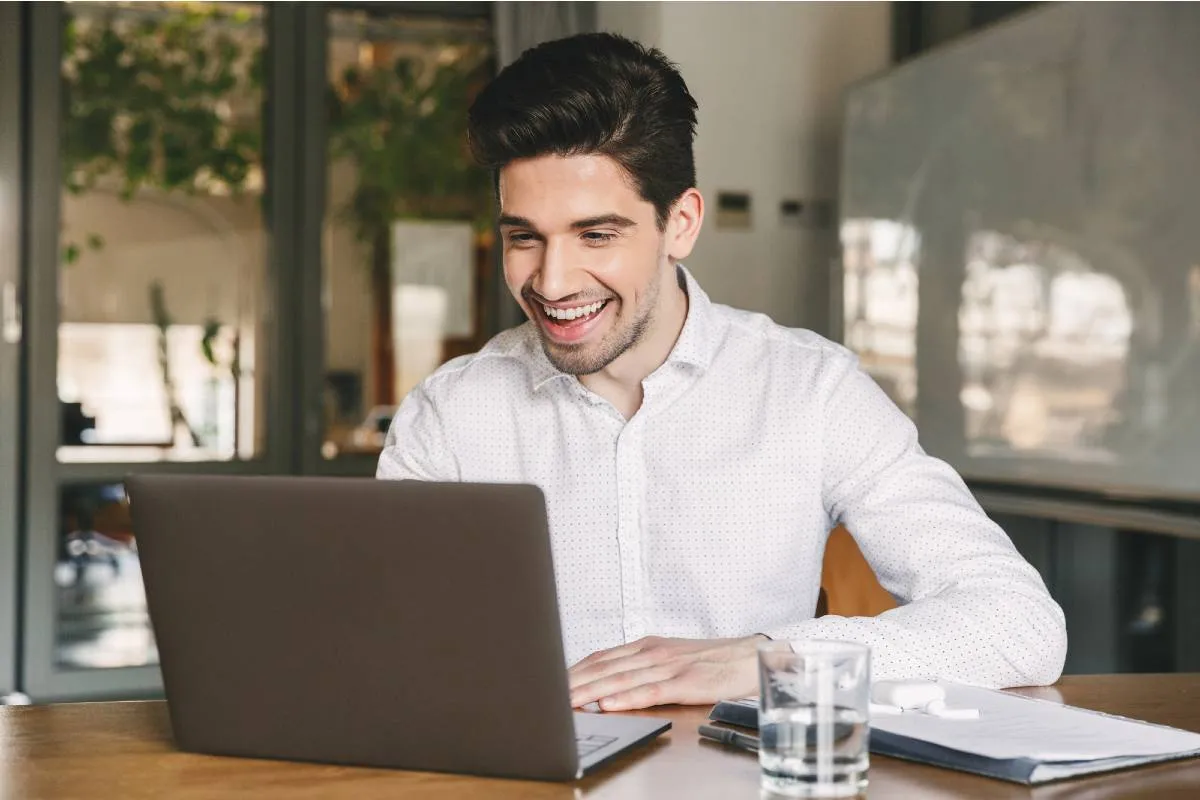 Imagem mostra um homem usando uma camisa branca em frente a um notebook. Ele está olhando para o computador e rindo. Tem um copo de água no canto da mesa.