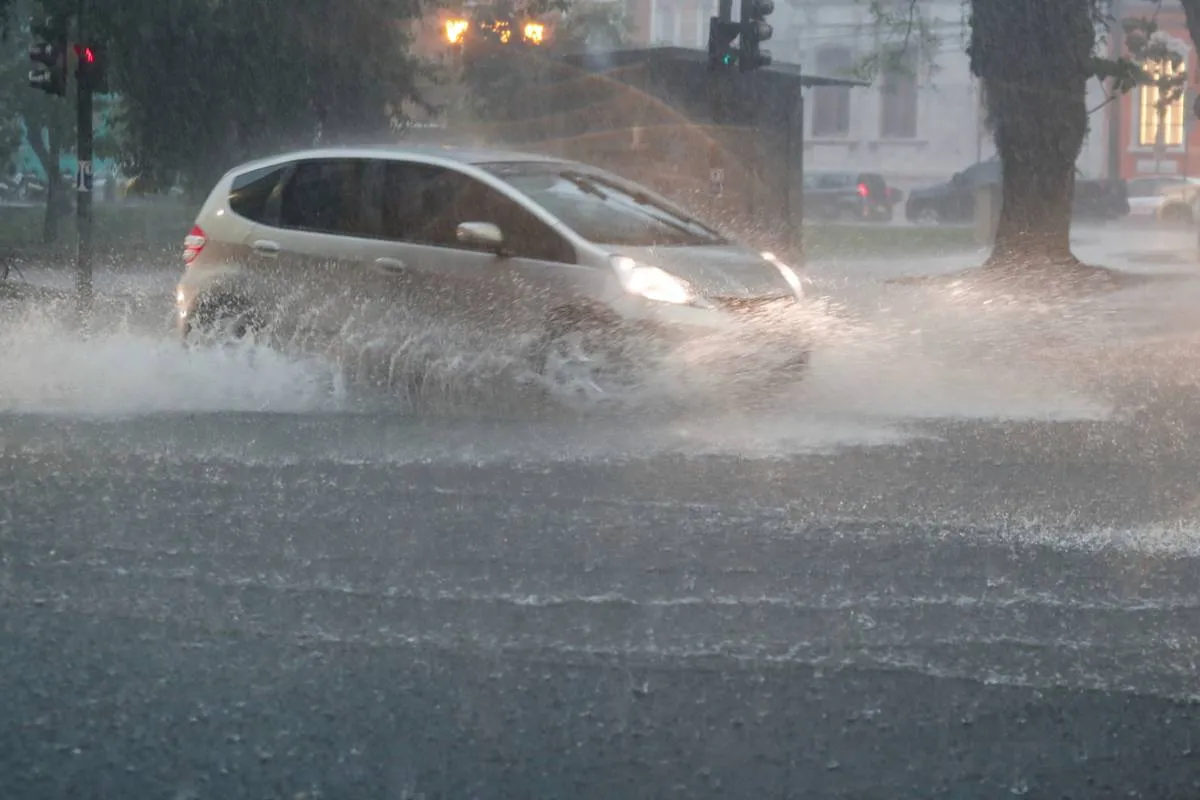 Imagem mostra um carro circulando em uma rua alagada. o veículo, um FIt cinza, está com a luz acesa e fazendo uma onda em um trecho alagado.