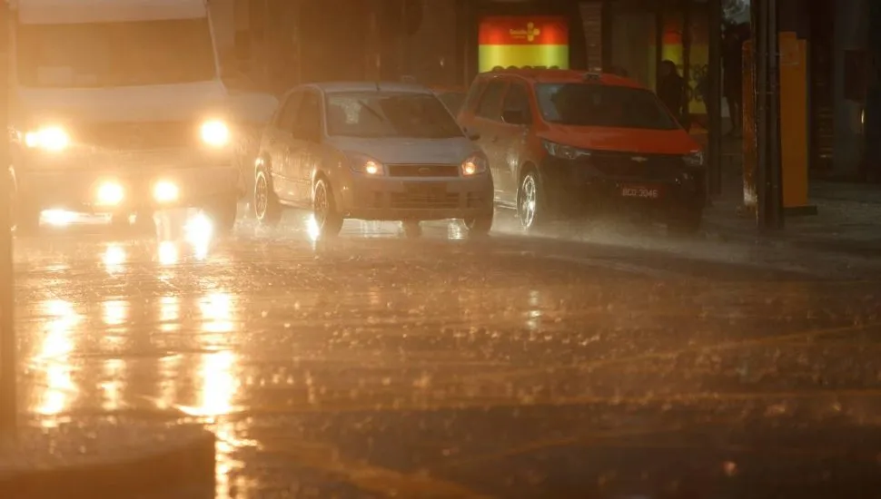 Imagem mostra carros parados em um semáforo durante um momento de forte temporal. Uma van branca e um carro cinza estão com as luzes ligadas. Um táxi está parado ao lado. Há muita chuva e os pingos caem fortes no asfalto.