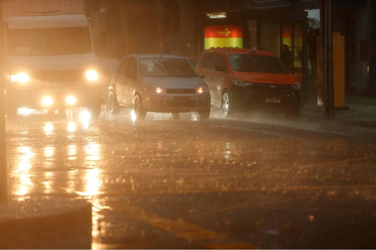 Imagem mostra carros parados em um semáforo durante um momento de forte temporal. Uma van branca e um carro cinza estão com as luzes ligadas. Um táxi está parado ao lado. Há muita chuva e os pingos caem fortes no asfalto.