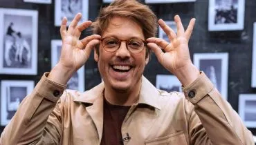 Imagem mostra Fabio Porchat. O humorista está na frente de um painel com várias fotos, usando uma jaqueta bege, sorrindo e segurando na ponta do seu óculos.