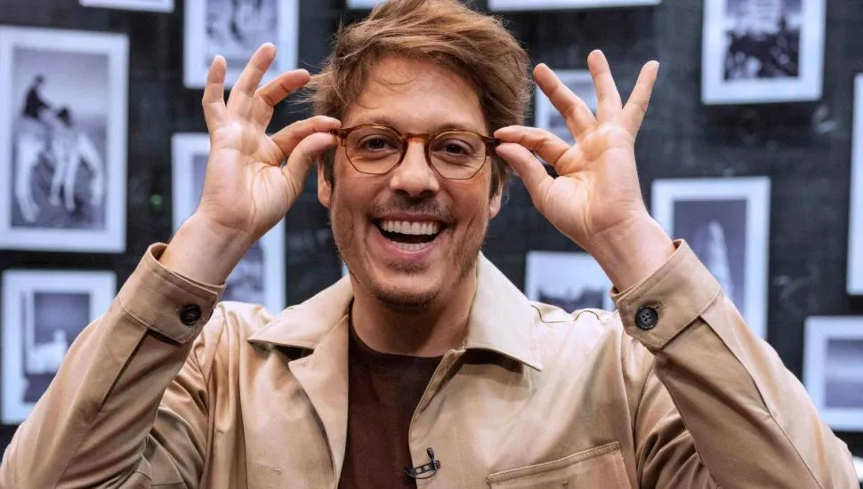 Imagem mostra Fabio Porchat. O humorista está na frente de um painel com várias fotos, usando uma jaqueta bege, sorrindo e segurando na ponta do seu óculos.