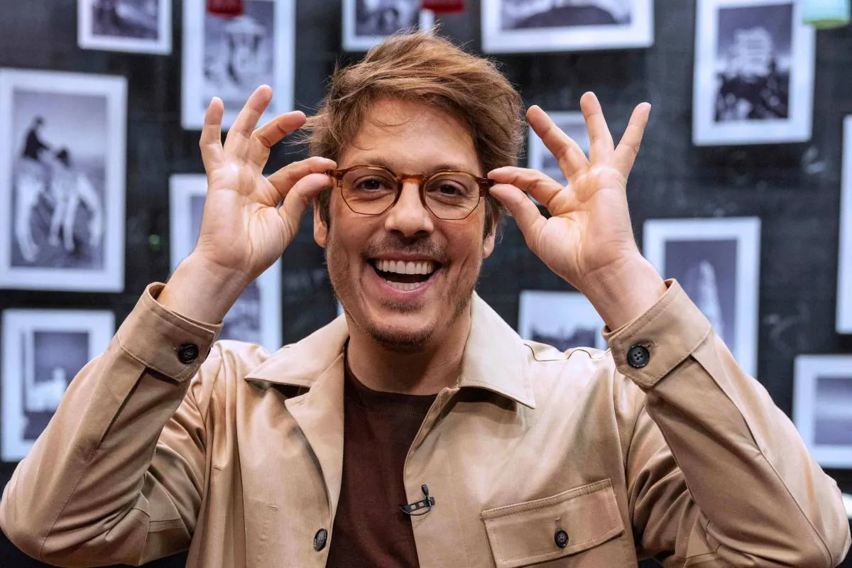 Imagem mostra Fabio Porchat. O humorista está na frente de um painel com várias fotos, usando uma jaqueta bege, sorrindo e segurando na ponta do seu óculos.