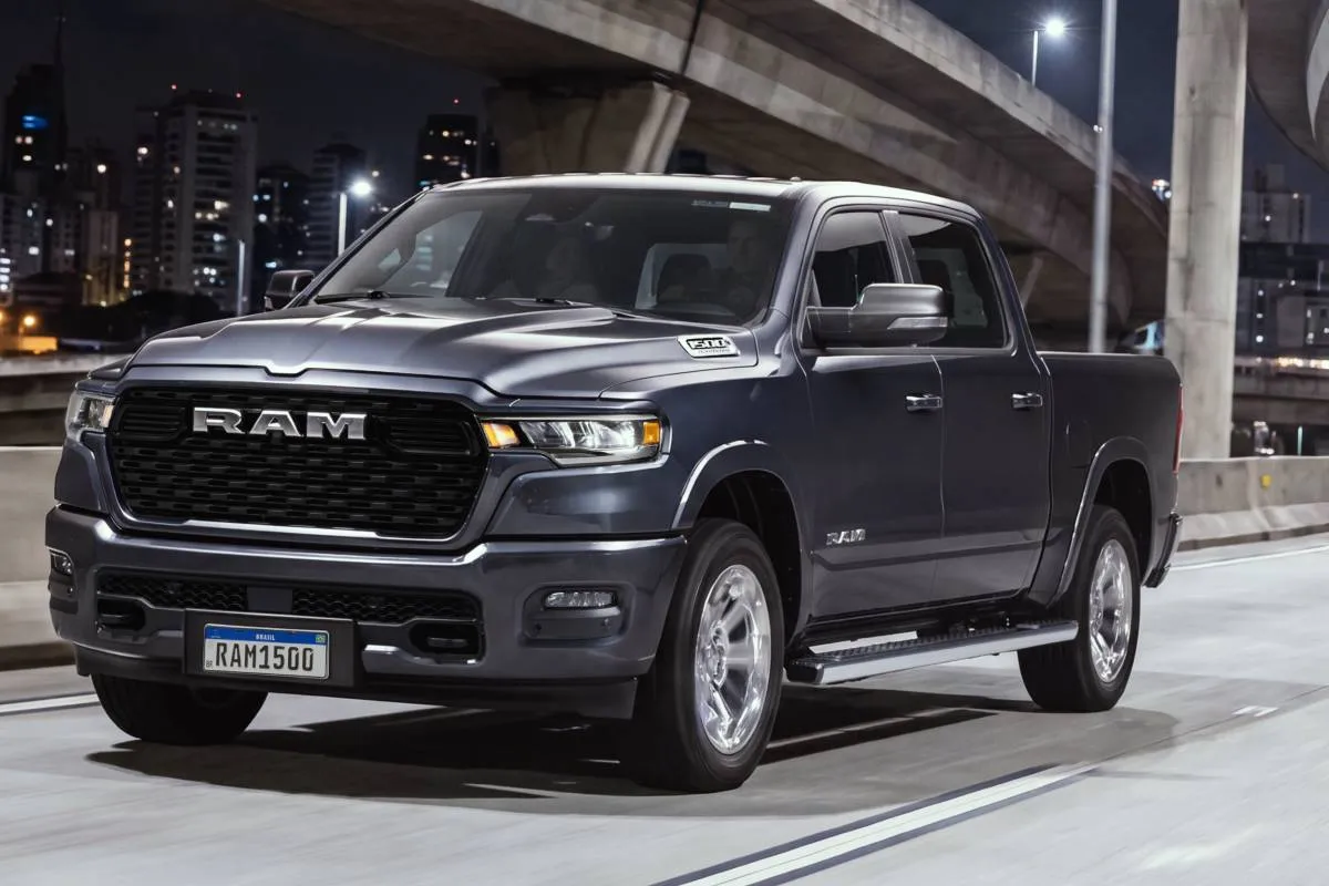 Imagem mostra a nova RAM 1500 big horn. No detalhe a frente da nova picape de grande porte da marca na cor azul marinho, com uma grade robusta escrito RAM em uma pista durante a noite.