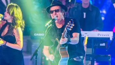 Imagem mostra a banda Blitz durante um show, com Evandro Mesquita com um violão cantando ao microfone e luzes verdes.