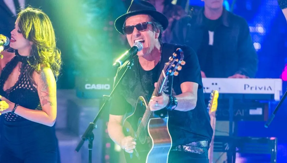 Imagem mostra a banda Blitz durante um show, com Evandro Mesquita com um violão cantando ao microfone e luzes verdes.