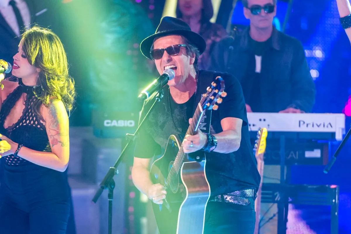 Imagem mostra a banda Blitz durante um show, com Evandro Mesquita com um violão cantando ao microfone e luzes verdes.