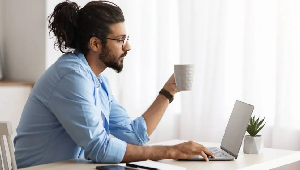 Imagem mostra um homem jogando no computador. Ele veste uma camisa azul, tem cabelo longo preso e está com um xícara branca.