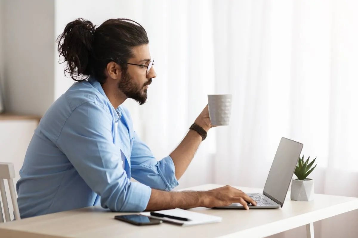 Imagem mostra um homem jogando no computador. Ele veste uma camisa azul, tem cabelo longo preso e está com um xícara branca.