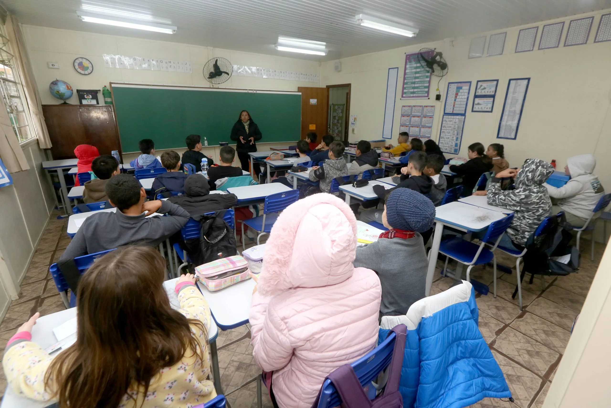 A imagem mostra uma sala de aula da Escola Municipal Vila Zanon. Os alunos estão de costas e a imagem mostra o quadro.