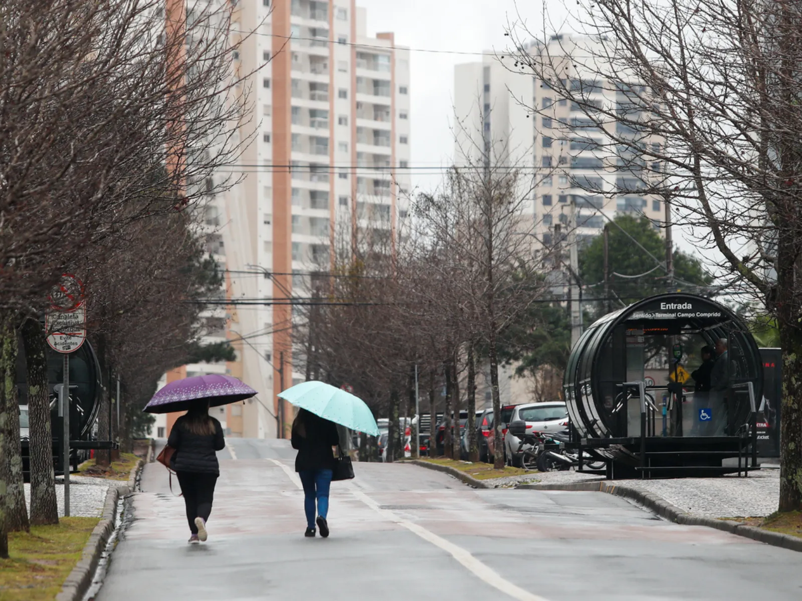 Fotografia de duas pessoas andando com guarda-chuvas na canaleta de ônibus em Curitiba.