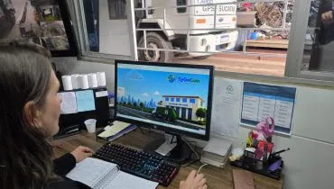 Reciclagem em Curitiba - mulher olhando sistema no computador e a sua frente um pátio de reciclagem com caminhão de sucatas