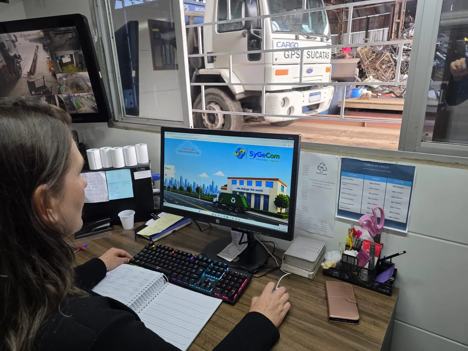 Reciclagem em Curitiba - mulher olhando sistema no computador e a sua frente um pátio de reciclagem com caminhão de sucatas